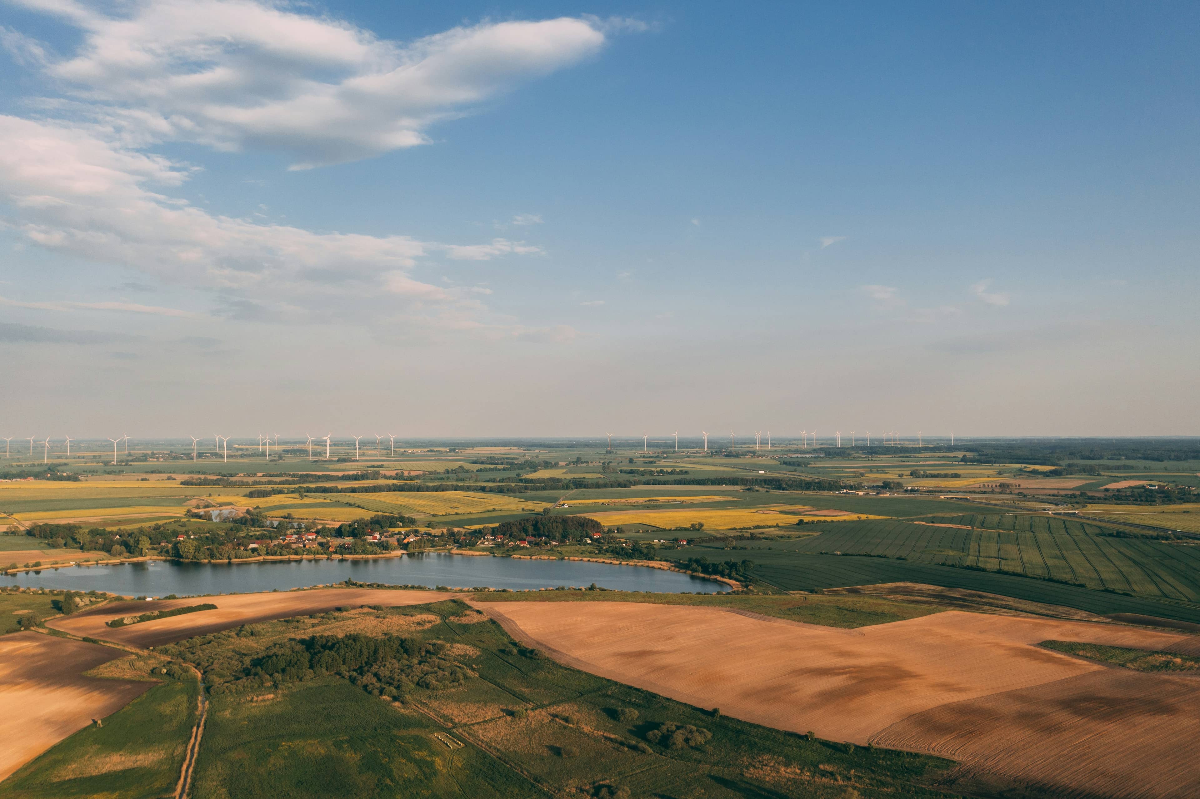 Aerial view of farmland with patchwork fields and wind turbines on the horizon under a blue sky.